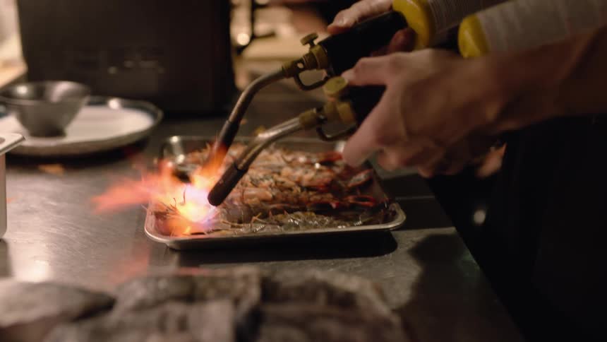 A chef expertly wields two powerful blowtorches searing a tray of large shrimp as bright orange flames lick across them in a professional kitchen setting