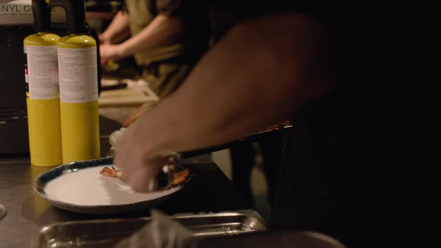 A chef carefully transfers grilled seafood from a metal tray onto a white plate in a dimly lit professional kitchen two yellow culinary torches stand on the stainless steel counter next to the plating area