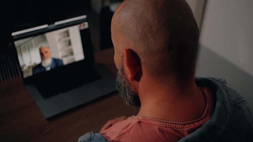 A man with a shaved head and gray beard is shown from behind as he participates in a video call on his laptop on the screen an older man with a white beard appears to be speaking during the virtual meeting