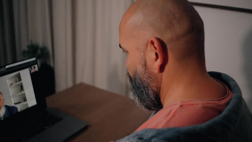A bald bearded man sits at a desk engaging in a video call on his laptop