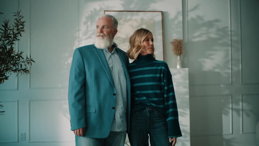 A distinguished man and a stylish woman stand close together posing as dappled light and shadows slowly shift and move across them and the tastefully decorated wall behind them