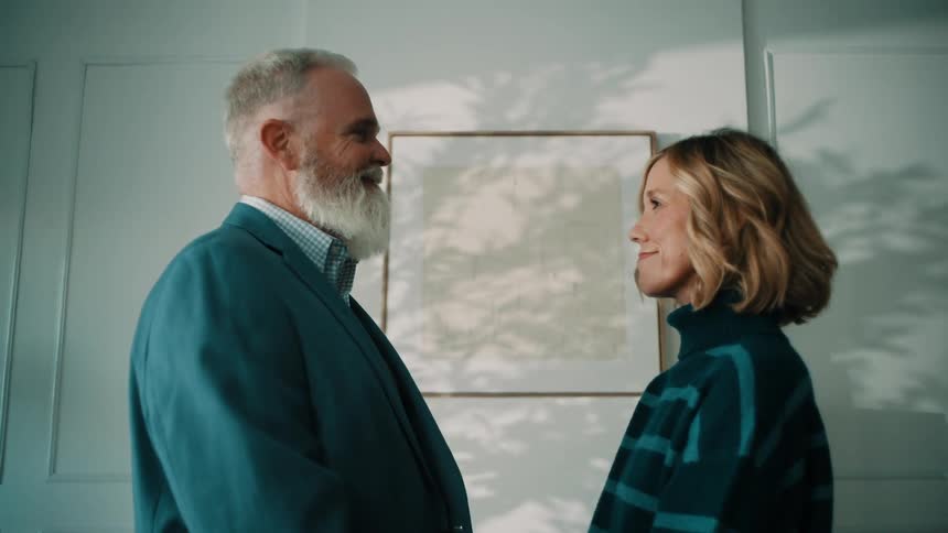 A middle aged couple a man with a white beard and a woman with short blonde hair stand facing each other and smiling affectionately in a well lit modern room