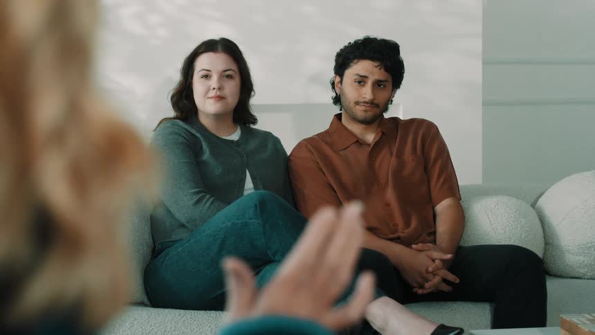 A man and a woman sit on a lightcolored sofa looking attentively toward someone in the foreground who is gesturing with their hand the scene is shot from a shallow depthoffield perspective focusing on the couples calm but engaged expressions during