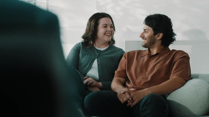 The man and woman sit closely on a sofa sharing a joyful and intimate moment as they look at each other and laugh the bright natural light from a window enhances the warm and genuine atmosphere of their interaction