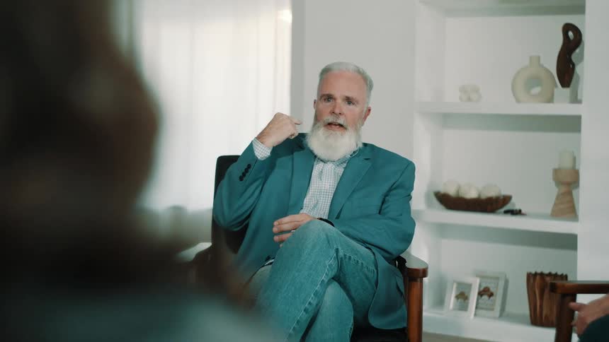 An older man with a white beard and a blue blazer sits in a chair and gestures while speaking during a group session