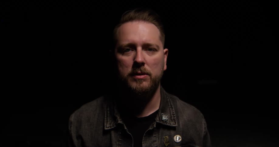 Bearded man wearing a dark jacket looks straight ahead under low lighting against a black background