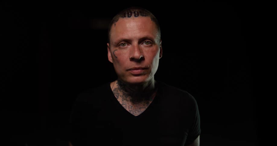 Man with facial and neck tattoos wearing a black shirt faces forward under low lighting against a black background