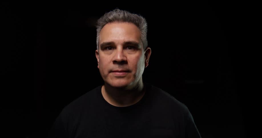 A man looks directly at the camera in a dark studio under dramatic lighting