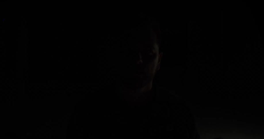 A young man stands inside a dark room as a brief flash of light reveals his face