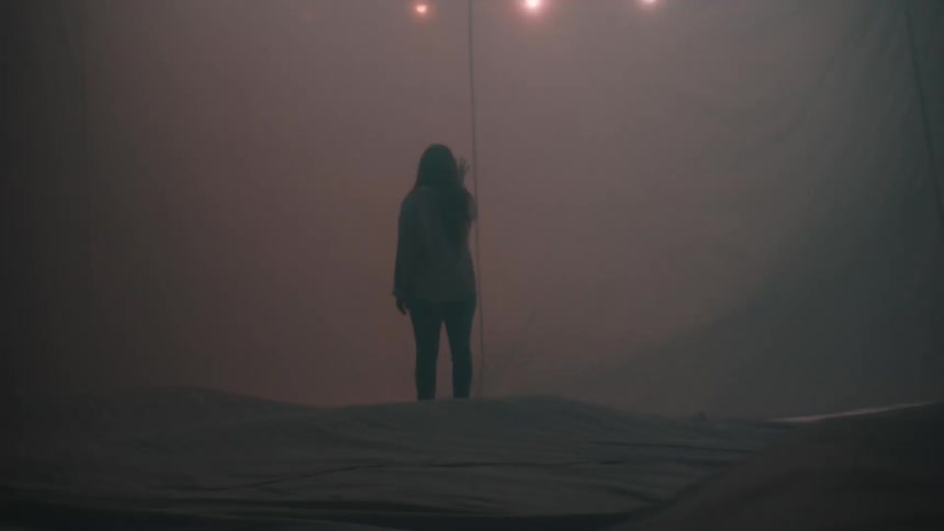 A lone person stands motionless in a fog filled space under soft warm lighting