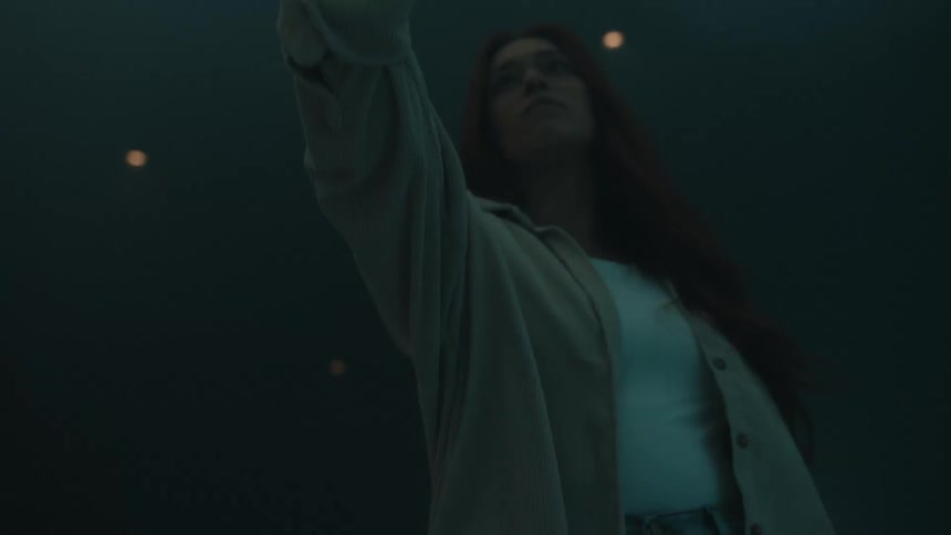 A woman with long red hair interacts with large draped white fabric in a misty atmospheric room the cinematic lighting shifts between warm orange glows and cool blue tones highlighting her movements and profile through thick fog