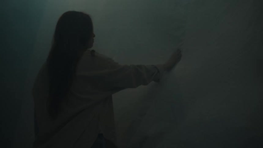 A woman stands with her back to the camera reaching out to touch a large semitransparent white sheet draped in a dark room the scene is shrouded in heavy mist or smoke with the lighting shifting dramatically from a warm orange glow to a cool blue hue