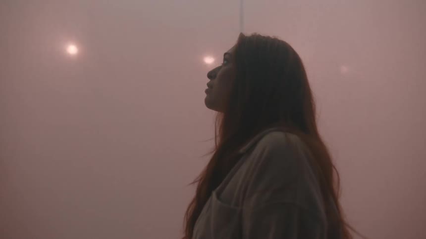 Young adult woman wearing a white shirt standing next to a white curtain with a nervous facial expression surrounded by strobing colored lights indoors