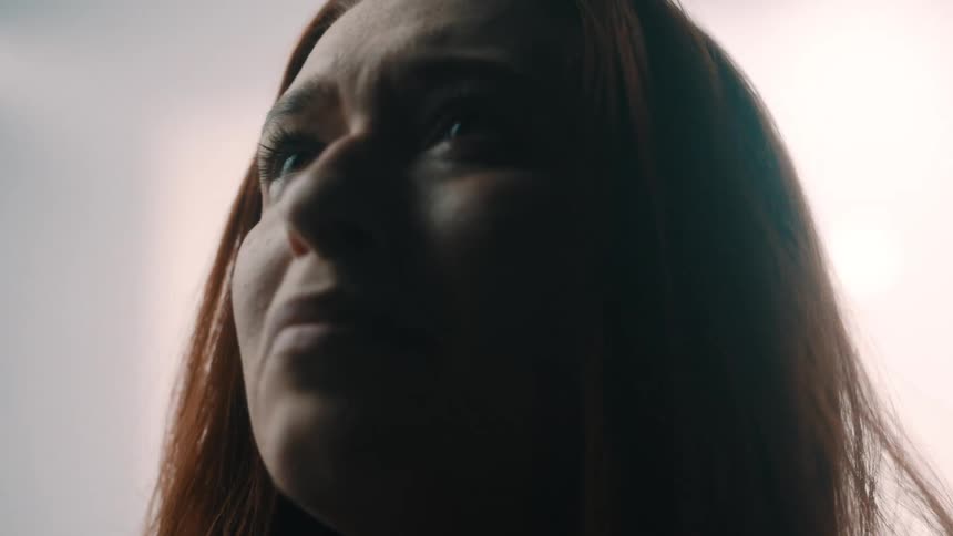 A womans pained expression softens into one of thoughtful hope as she lowers her hands from her hair and gazes upward into a bright ambiguous light