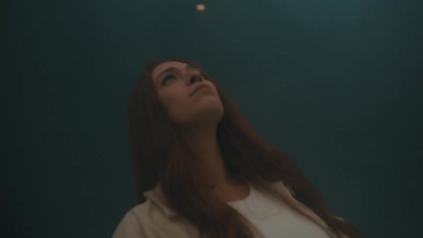 A young woman with long red hair wearing a white shirt and tan jacket looks upward as fog and light move around her against a dark background