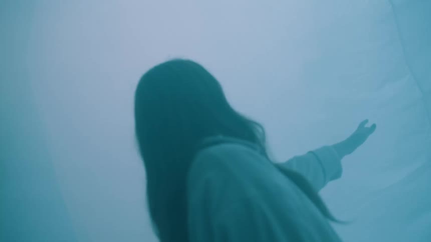 A woman with long dark hair reaches out toward a large white fabric sheet her movements obscured by thick fog and shifting cinematic lighting the scenes transition between warm orange glares moody teal shadows and abstract closeups of light reflectin