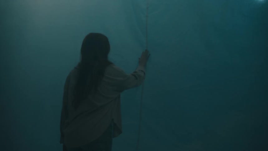 In a hazy dimly lit setting a woman with long dark hair stands with her back to the camera reaching out to touch a large pale curtain that is illuminated by shifting colored lights