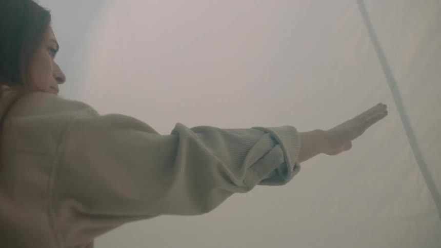 In a misty setting a woman reaches out to touch a large white fabric sheet that is backlit by soft light the atmosphere shifts between warm hazy tones and a moody cool blue illumination as she interacts with the draped material