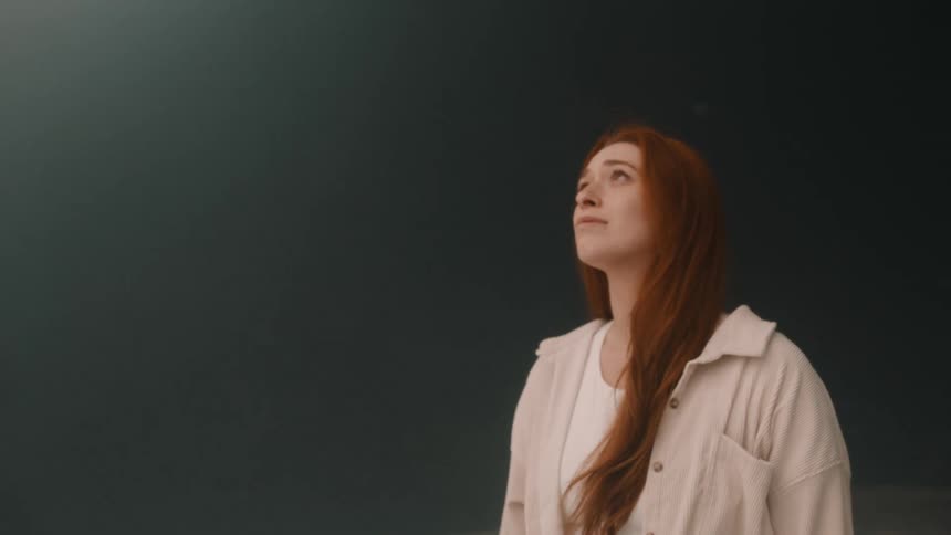 A young woman with long red hair and a white corduroy shirt looks upward against a dark plain background