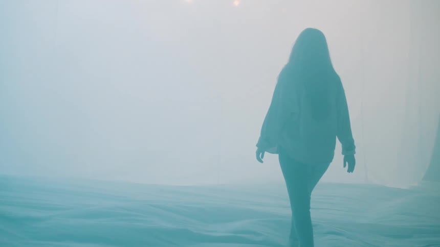 A woman walks amidst windblown fabric illuminated with changing color light