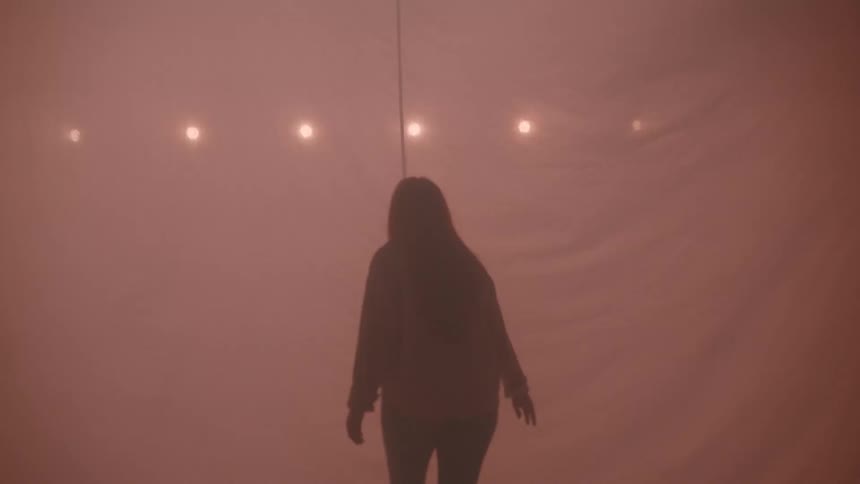 A young woman with long dark hair is seen in profile looking upward as flashing lights illuminate a large thin fabric sheet behind her in a dark space