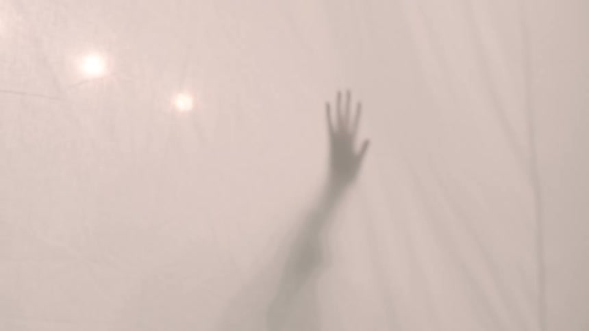 The shadow of a hand and arm reaching up is cast against sheer light colored fabric with a few blurry bright lights visible near the top
