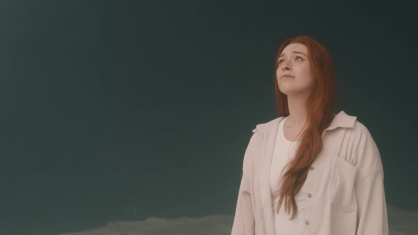 A woman with long red hair wearing a white shirt and cream jacket looks upward with a calm expression against a dark teal background