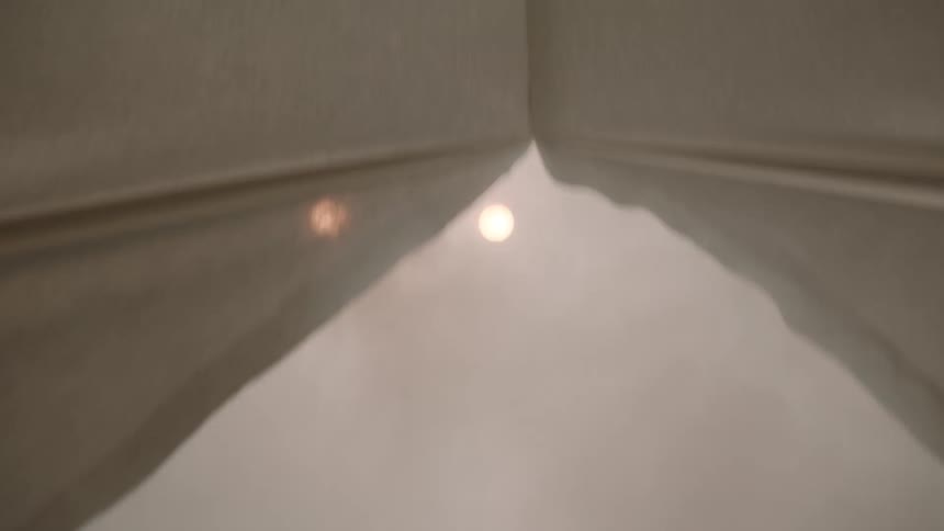 A pair of beige curtains slowly pulls apart to reveal bright outdoor light with a string of glowing blurry lights visible through the opening