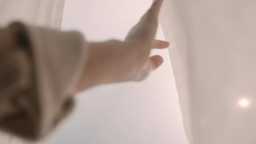 Hand of a woman opening white curtains revealing morning sunlight