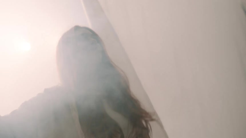 A woman pushes aside a fabric curtain and enters a foggy indoor space