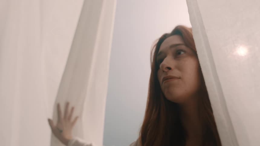 Backlit by a bright window a woman parts white curtains and enters a darker room her amber hair catching the light and flowing as she walks purposefully forward