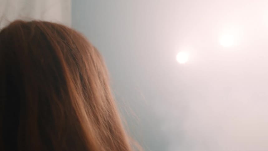 A hand opens sheer white curtains to a bright backlit sky before a woman walks through them and the curtains close again
