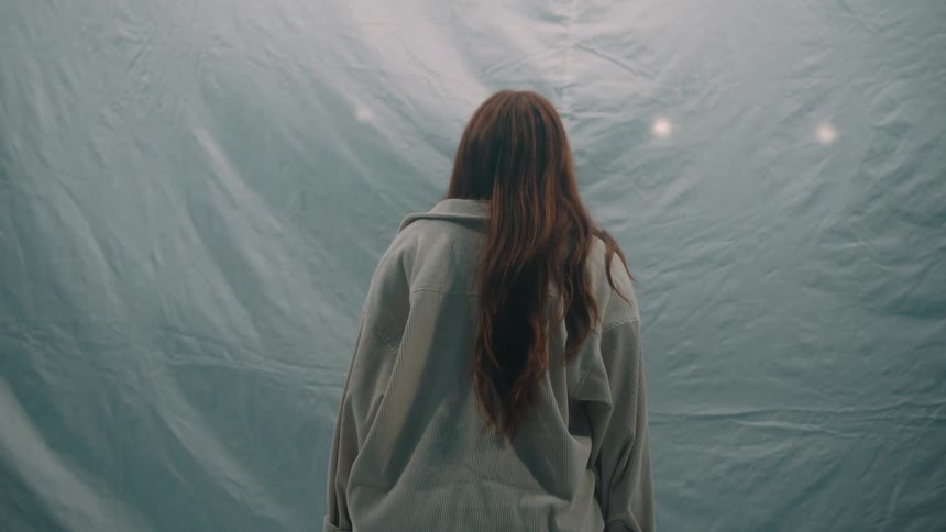 A view from behind shows a woman with long red hair wearing a tan shirt standing in front of a large wrinkled white fabric backdrop