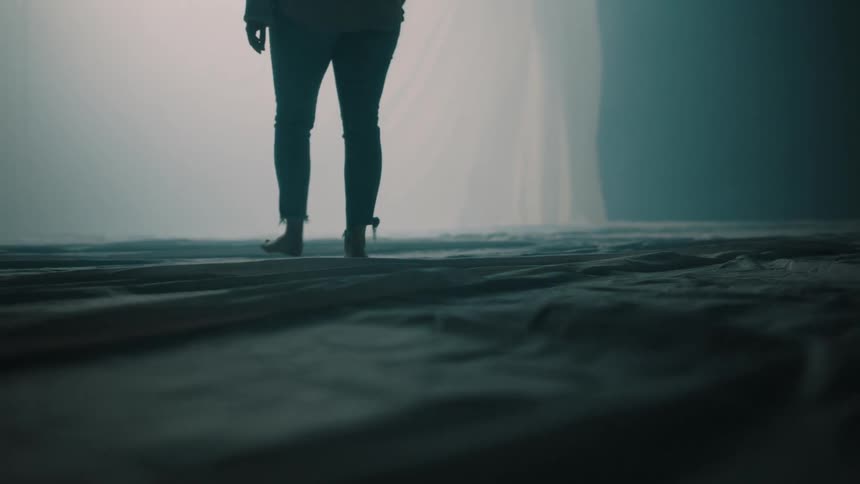 A young woman walks across a stretch of fabric inside a large tent moving toward a section ahead thats illuminated by bright light