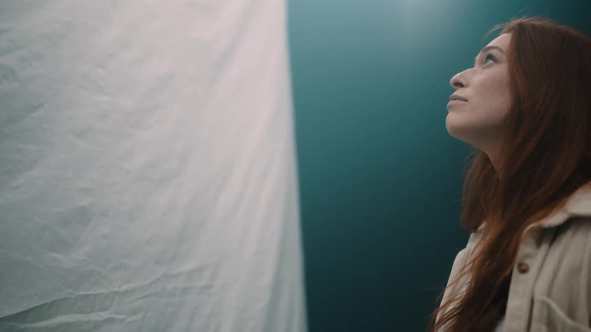 A woman with long red hair gazes upward with a serene expression toward a bright light source she is positioned on the right side of the frame against a teal wall and a draped white cloth