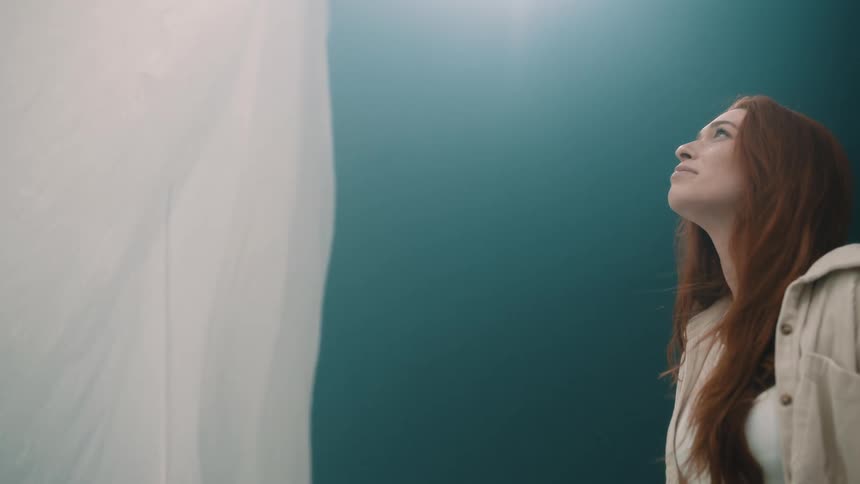 A woman gazes upward with an expression of serene wonder her face illuminated by a soft overhead light as a large white fabric structure billows gently beside her