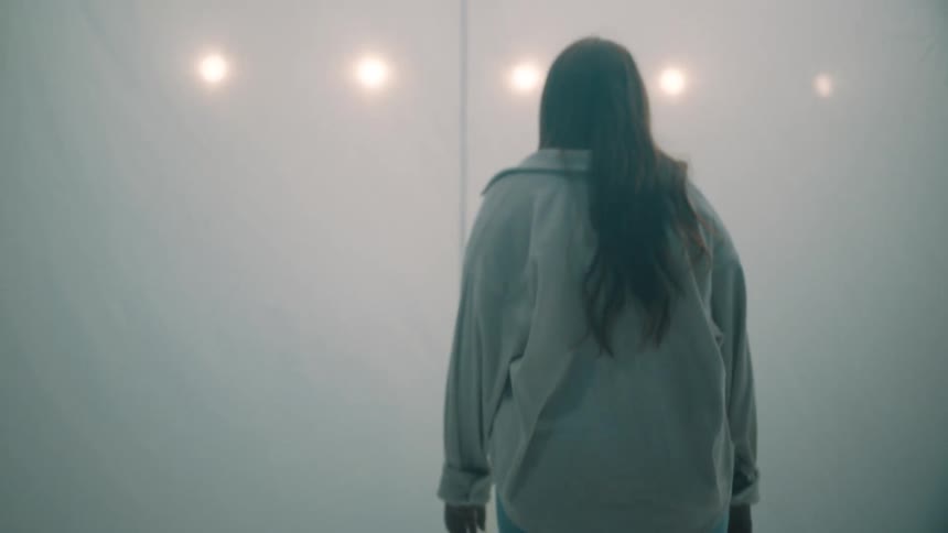 Young adult woman wearing a shirt and jeans walking towards a white curtain with a row of spotlights seen through the material indoors
