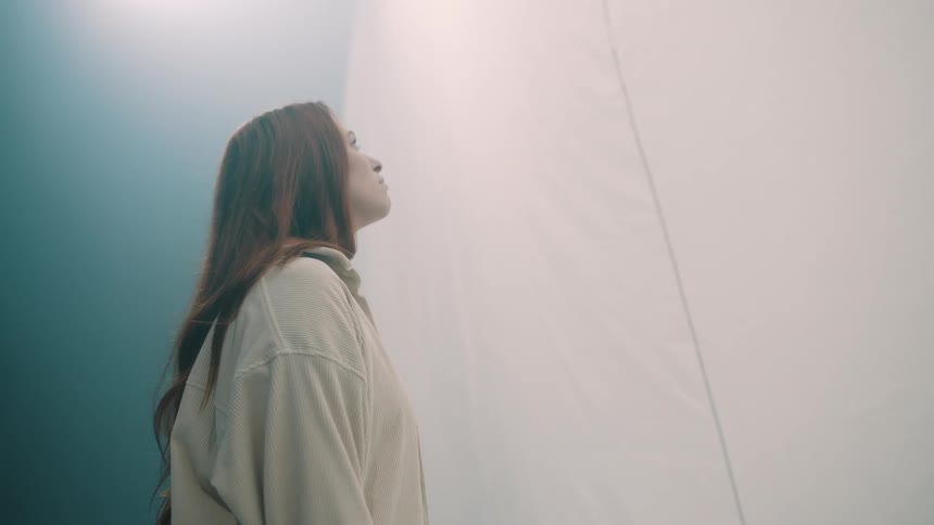 A young woman stands inside a bright room her long hair falling freely as she gazes upward toward a fabric wall in front of her bright light reflecting softly across its surface