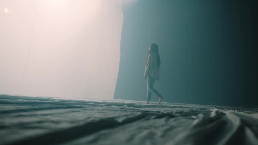 A woman walks across a cloth covered floor toward bright diffused light in a dim studio