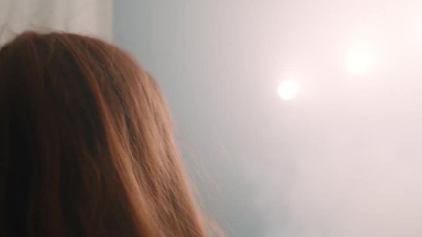 A view from behind shows a woman with long red hair wearing a tan shirt standing in front of two large white fabric sheets that she begins to pull apart to reveal a bright light