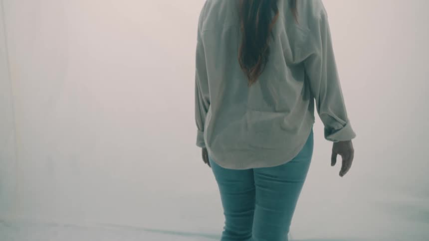 Following from behind a barefoot woman walks slowly across a rumpled white surface venturing into a bright hazy and ambiguous space as the camera tilts up to follow her