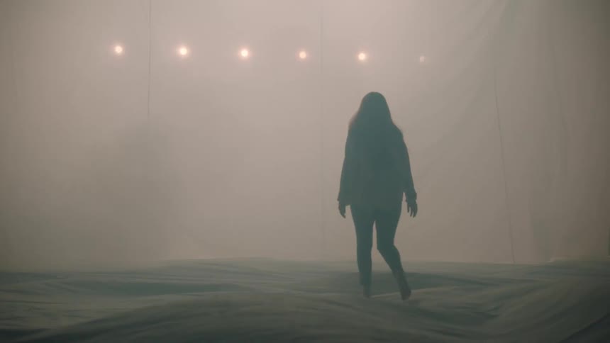 The video depicts a woman in silhouette walking through a dimly lit fogfilled room toward a row of glowing lights as she reaches the far side she extends her hand to touch a translucent hanging fabric sheet
