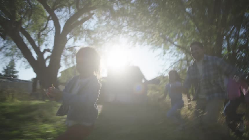 A man and three young children run joyfully through a sundrenched backyard during the day moving across a grassy area with trees and a house visible in the background as sunlight filters through the leaves