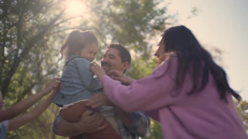 Happy family of four hugs and tickles each other outside with sunlight through green trees