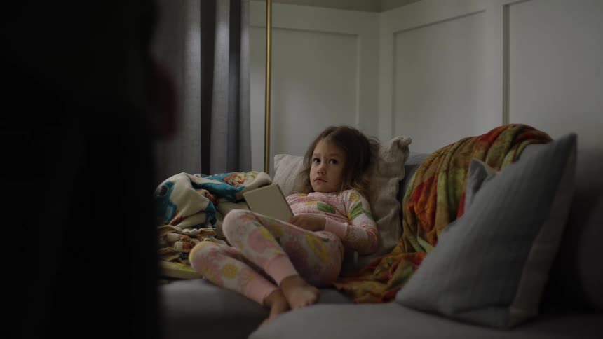 Young girl wearing pink pajamas and reading a book while occasionally looking at another person standing in front of her indoors during night time