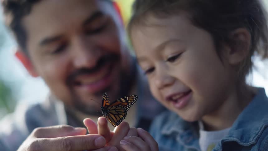 Father and child observe a butterfly resting between their fingers outdoors