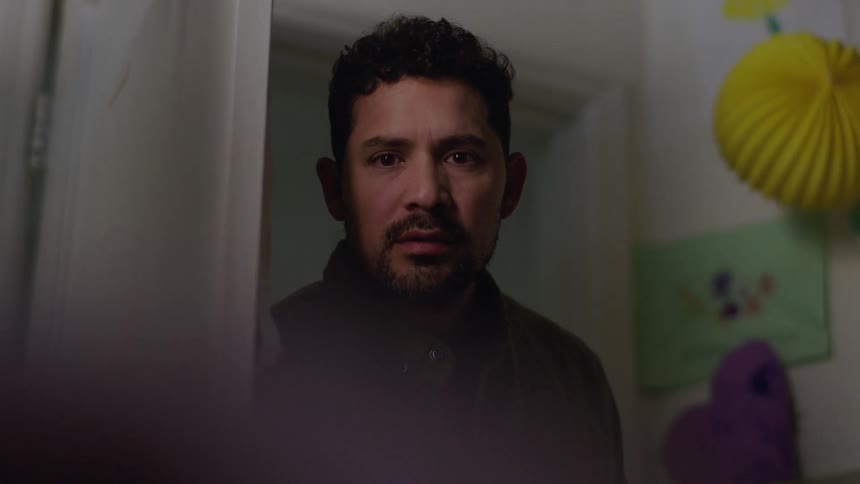 A close up shot shows a man with dark curly hair and a beard as he slowly opens a door and looks into a room with a serious expression