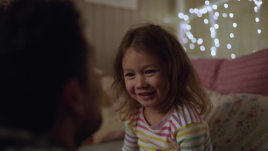 A young girl in a striped shirt sits on a bed looking attentively at a man whose back is to the cameraas the man speaks her serious expression quickly transforms into a wide joyful laugh