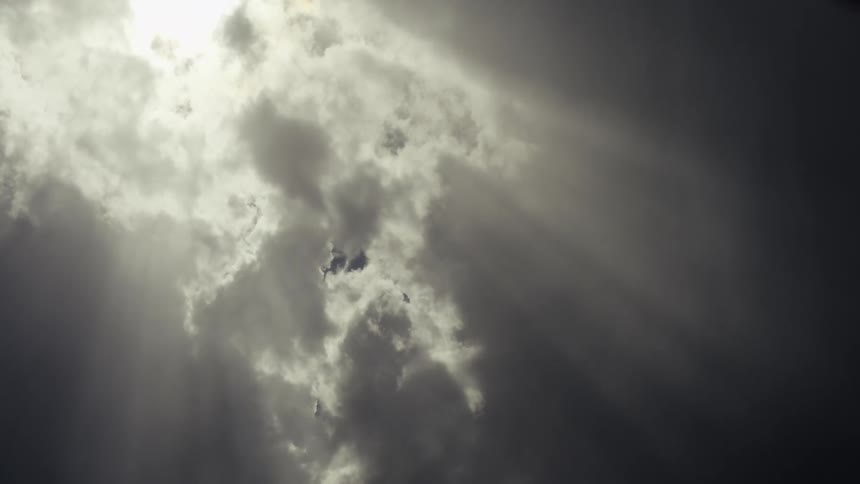Bright sunbeams break through a thick layer of moving clouds creating a dramatic contrast between the brilliant white light and the darker shadowed portions of the sky as the sun remains partially obscured