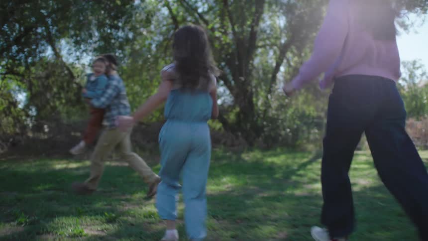 A happy family of four runs through a sundrenched grassy field toward a wooded area the parents playfully catch and swing their two young daughters as they all laugh together in the sunlight